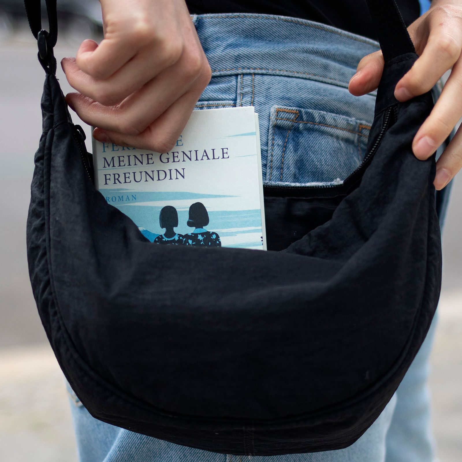 Foto einer Umhängetasche, aus der eine Hand das Buch "Meine geniale Freundin" von Elena Ferrante hochhebt, in der Ausgabe der Reihe Suhrkamp Pocket