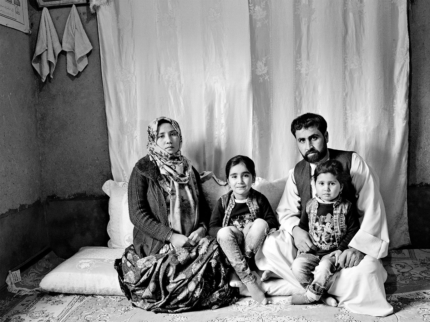 Eine vierköpfige afghanische Familie, Mutter, Vater und zwei Kinder, die auf dem Boden eines Hauses sitzen. Der Vater hat ein Kind auf dem Schoß. © Kaveh Rostamkhani