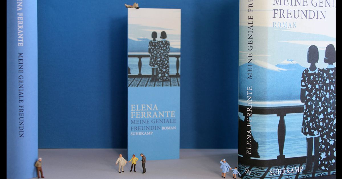 Links und rechts steht das aufgestellte Buch »Meine Geniale Freundin«, einmal mit und einmal ohne Schutzumschlag. In der Mitte ist ein aufgestelltes Lesezeichen vor dem Miniaturfiguren stehen. 