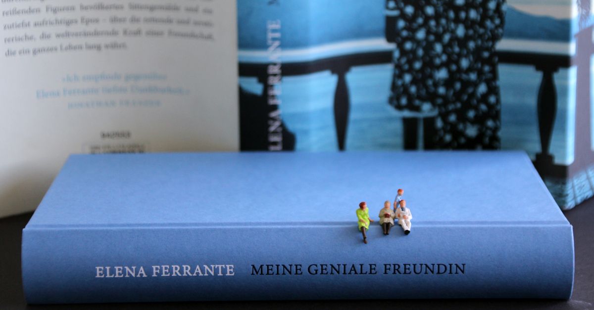 Liegendes Buch »Meine Geniale Freundin« ohne Schutzumschlag, auf dem drei Miniaturfiguren sitzen. Im Hintergrund ist der aufgeklappte Schutzumschlag aufgestellt. 